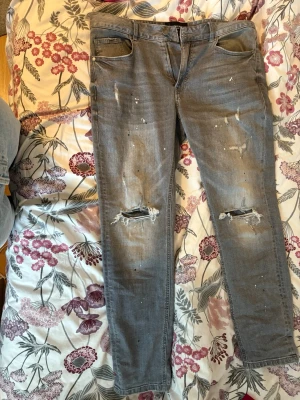 Grå slitna skinny jeans med hål - Säljer ett par grå skinny jeans med slitningar och hål på knäna. Jeansen har en tvättad look, färgstänk och klassiska fem fickor. Perfekta för en avslappnad och trendig stil.