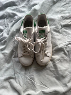 Adidas Stan Smith vita sneakers - Klassiska vita Adidas Stan Smith sneakers med gröna detaljer på hälen och tungan. Skorna har perforerade ränder på sidorna, vita snören och är tillverkade i slätt läder. Perfekta för en clean och tidlös stil.