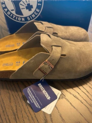  Birkenstock boston grey - Säljer ett par snygga gråa tofflor från Birkenstock i mjuk mocka. Modellen har en bred rem med metallspänne och klassisk korksula för extra komfort. Perfekta för dig som gillar stilrena och bekväma skor.