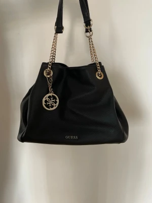 Äkta Guess svart handväska / tote bag med gulddetaljer - Säljer en jättefin och 100% äkta Guess handväska i mjukt, svart konstläder med eleganta guldkedjor och tillhörande Guess-logohänge. Modellen heter Isabeau Fluid och är både rymlig och väldigt stilren.