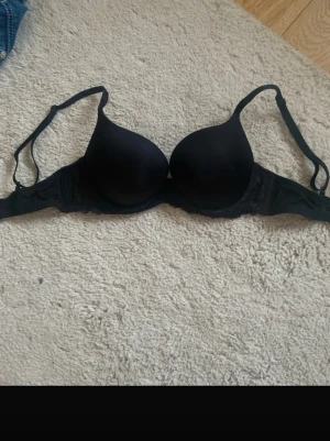 Svart push-up bh med spetsdetaljer - Snygg svart push-up bh med vadderade kupor och justerbara axelband. Sidorna har fina spetsdetaljer som ger en extra touch.  70b