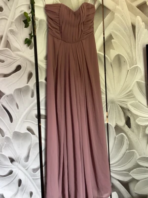 Lång axelbandslös lila klänning - Elegant långklänning i en mjuk lila ton med axelbandslös design och veckad överdel. Klänningen har en flytande kjol och dragkedja i ryggen. Perfekt för speciella tillfällen där du vill känna dig extra fin.