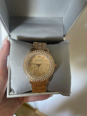 Guldig klocka Marc by Marc Jacobs - Snygg guldig klocka från Marc by Marc Jacobs med rund urtavla och gnistrande stenar runt hela boetten. Länken är i metall och urtavlan har märkets logga i bokstäver. Perfekt accessoar för att lyfta din stil. Vet inte om den funkar men de är nog bara att sätta i ett nytt batteri💕den är sparsamt använd men lite slitage eller tecken på användning finns. Har tyvärr inte kvittot kvar🤗