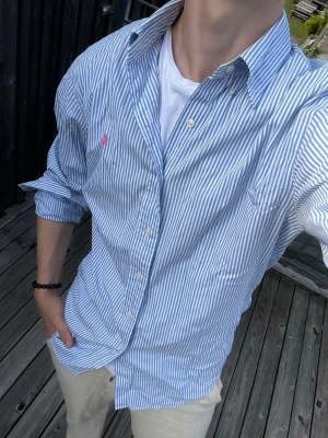 Blårandig skjorta från Ralph Lauren - Säljer en klassisk blårandig skjorta från Ralph Lauren i modellen Custom Fit. Skjortan har vita knappar, lång ärm och den ikoniska rosa loggan broderad på bröstet. Tillverkad i mjuk bomull och passar perfekt till en avslappnad men stilren look.