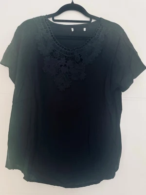 Svart blus med spetsdetaljer - Svart blus med rund halsringning och snygga spetsdetaljer framtill. Blusen har korta ärmar och ett luftigt, mjukt tyg som ger en avslappnad men ändå stilren look. Perfekt att matcha med jeans eller kjol för en trendig outfit.
