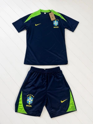 Brasilien Nike fotbollsställ blå/grön - Snyggt fotbollsställ från Nike i Brasiliens färger. Setet består av en mörkblå t-shirt med gröna axelpartier och gula detaljer samt matchande shorts. Både tröja och shorts har Brasilien-logga och Nike swoosh. Perfekt för träning eller match!