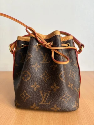 Louis Vuitton Monogram Noé BB väska - Snygg brun handväska från Louis Vuitton i klassiskt monogrammönster. Modellen är Noé BB med justerbar axelrem i ljust läder och dragskostängning. Väskan har detaljer i guld och röda kanter. Perfekt för dig som gillar lyxiga accessoarer.