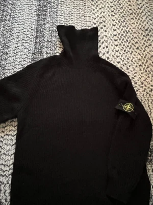 Svart polotröja från Stone Island - Stickad svart polotröja från Stone Island med klassisk patch på ärmen. Tröjan har ribbad struktur, hög polokrage och lång ärm. Perfekt för dig som gillar streetwear och vill ha något både stilrent och bekvämt.