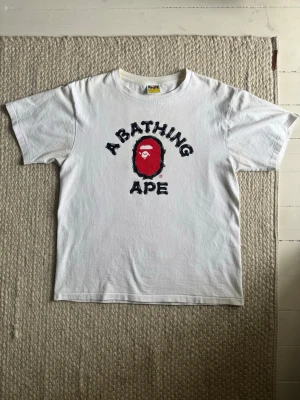 Vit Bape t-shirt med tryck - Säljer en klassisk vit t-shirt från Bape med det ikoniska apansiktet och texten 'A Bathing Ape' i svart och rött på bröstet. Baksidan har en liten 'Bape'-logga i nacken. Skön passform och perfekt till streetwear-stilen.
