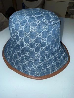 Gucci hat - Snygg bucket hat från Gucci i blå denim med klassiskt GG-mönster över hela hatten. Hatten har en brun läderkant runt brättet som ger en exklusiv touch. Perfekt accessoar för att lyfta din outfit. Går ner i pris vid snabbaffär