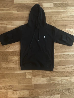 Svart hoodie  - Svart hoodie med huva och dragsko. Liten vit logga broderad på bröstet. Klassisk känguruficka framtill. Perfekt för en avslappnad och stilren look.den är 62 centimeter lång