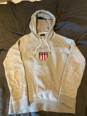 Grå hoodie från GANT med emblem - Snygg grå hoodie från GANT med broderat emblem på bröstet i rött, vitt och blått samt vita dragsnören i huvan. Klassisk passform och mjukt material, perfekt för en avslappnad stil. GANT-logga diskret broderad ovanför emblemet.