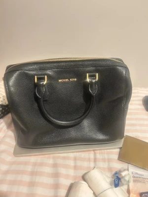 Svart handväska från Michael Kors - Snygg svart handväska från Michael Kors i strukturerat läder med guldfärgade detaljer och dubbla handtag. Väskan har en klassisk, boxig form och dragkedja upptill. Perfekt för dig som vill ha en stilren och tidlös accessoar. Aldrig använt från mig! Fick den som en present men tror att det kanske har använts några gånger men i super super bra skick 