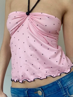 Rosa prickig bandeau topp med knyt - Söt rosa bandeau topp med svarta prickar och halterneck-band. Toppen har en snygg knytning i ryggen och vågig kant nertill. Perfekt för dig som gillar en feminin och trendig stil.