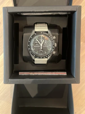 Omega x Swatch Speedmaster klocka - Säljer en Omega x Swatch Speedmaster klocka med svart urtavla och grått band. Klockan har kronograf, tachymeter och tydliga vita markeringar. Snygg och modern design, perfekt för dig som gillar stilrena accessoarer.