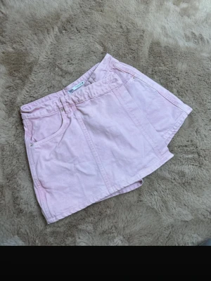 Ljusrosa denimskort med fickor - Supersöt ljusrosa skort i denim med klassisk femficksdesign och knapp framtill. Kjol framtill och shorts baktill ger en avslappnad men trendig look. Perfekt för dig som vill ha både stil och komfort i ett plagg.