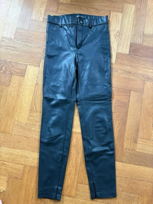 Svarta leggings i fuskläder med dragkedja - Snygga svarta leggings i fuskläder med hög midja och bälteshällor. Tight passform och dragkedjor vid bensluten för extra detalj. Perfekta för en trendig look och lätta att matcha med olika stilar.