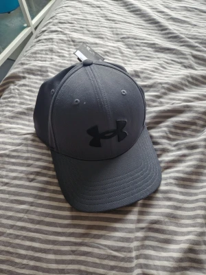 Mörkgrå keps från Under Armour - Snygg mörkgrå keps från Under Armour med böjd skärm och broderad svart logga framtill. Kepsen har klassisk sexpanelsdesign och justerbar passform. Perfekt accessoar för en sportig och stilren look.