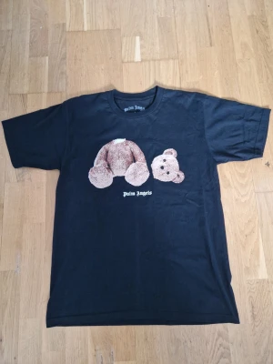 Svart Palm Angels t-shirt med björn - Svart t-shirt från Palm Angels med ikoniskt nalletryck på bröstet och logga under. Klassisk rund hals och korta ärmar. Perfekt för dig som gillar streetwear och vill sticka ut med en unik design.