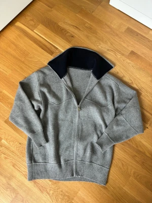 Grå stickad zip tröja  - Snygg grå stickad ziptröja med hel dragkedja och hög krage i mörkblått foder. Tröjan har ribbade muddar och nederkant, samt en avslappnad passform. Perfekt för lager-på-lager och chill dagar.