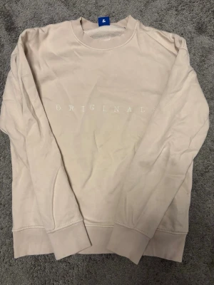 Beige sweatshirt med tryck - En stilren beige sweatshirt med texten 'ORIGINALS' tryckt framtill. Tröjan har rund halsringning, långa ärmar och ribbade muddar vid ärmslut och nederkant. Perfekt för en avslappnad och trendig look.