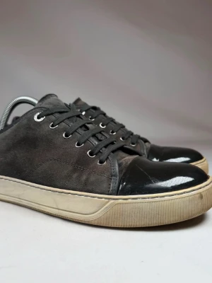 Svarta sneakers med lackad tå - Snygga svarta sneakers med ovandel i mocka och glansig lackad tå. Skorna har svarta skosnören, silverfärgade öljetter och en vit sula med beige inslag. Perfekta för dig som gillar en stilren men ändå unik look.
