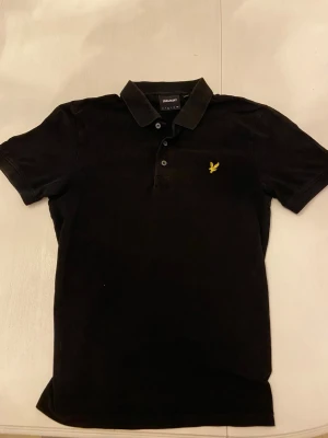 Svart pikétröja från Lyle & Scott - Klassisk svart pikétröja från Lyle & Scott med gul broderad logga på bröstet. Tröjan har krage, knappar framtill och korta ärmar. Perfekt till både jeans och shorts för en stilren look.