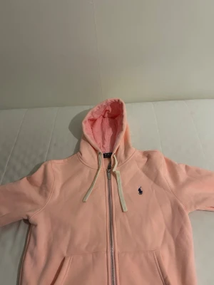 Ljusrosa hoodie med dragkedja - Säljer en ljusrosa hoodie med dragkedja och huva. Hoodien har vita snören, fickor framtill och en liten broderad logga på bröstet. Perfekt för en chill och avslappnad stil.