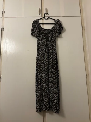Svart blommig långklänning - Snygg svart långklänning med vita blommor och korta puffärmar. Klänningen har en smockad överdel och söt offshoulder-ringning. Perfekt för dig som gillar romantisk och trendig stil.