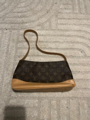 Brun monogram axelremsväska - Snygg axelremsväska i klassiskt brunt monogrammönster med beige läderdetaljer och lång rem. Väskan har en avlång form och är perfekt för dig som gillar stilrena accessoarer med lyxig känsla. Materialet är en mix av canvas och läder.