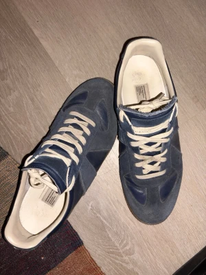 Blå sneakers med mockadetaljer - Säljer ett par blå sneakers med vita snören och beige innersula. Skorna har en kombination av mocka och läder på ovandelen samt en klassisk gummisula. Snygg retrostil som passar till jeans eller streetwear.