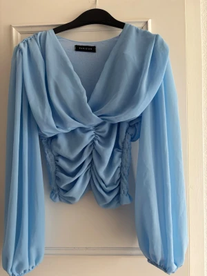 Ljusblå draperad blus från Parisian - Trendig ljusblå blus från Parisian med draperad framsida och rynkade detaljer. Blusen har långa, luftiga ärmar och smockad rygg för en bekväm passform. Tillverkad i ett tunt, flowy material som ger en elegant look.