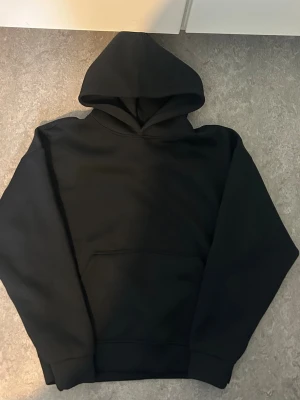 Svart basic hoodie med huva - En enkel och stilren svart hoodie med stor huva och magficka. Hoodien har ribbade muddar vid ärmar och nederkant. Perfekt för en avslappnad och casual look. Materialet ser ut att vara mjukt och bekvämt, troligen bomullsblandning.