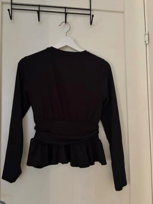 Svart långärmad topp med volang - Säljer en svart långärmad topp med markerad midja och volang nedtill. Toppen har en klassisk rund halsringning och är enkel att matcha till olika outfits. Perfekt för dig som gillar stilrena plagg med en twist.