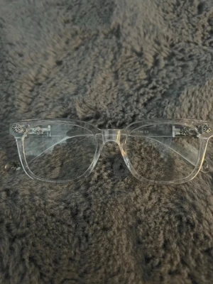 Transparenta Chrome Hearts Glasögon  - Snygga transparenta glasögon med rektangulär form och coola silverfärgade blommönster på bågarna. Glasögonen har en modern vibe och passar perfekt för dig som vill sticka ut lite extra. Tillverkade i plast med metalliska detaljer på sidorna.