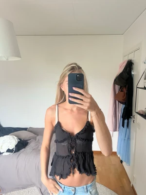 Svart volangig linnetopp med snörning - Säljer en svart linnetopp med tunna axelband, volangdetaljer och snörning framtill. Toppen är croppad och har en feminin, bohemisk vibe som passar perfekt till jeans eller kjol.