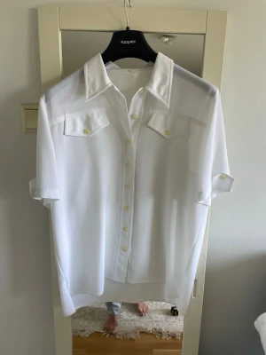 Vit skjorta med kort ärm och fickor - Stilren vit skjorta med korta ärmar och klassisk krage. Skjortan har två bröstfickor med lock och knappar framtill. Perfekt för en fräsch och enkel look. Tillverkad i ett lätt och luftigt material