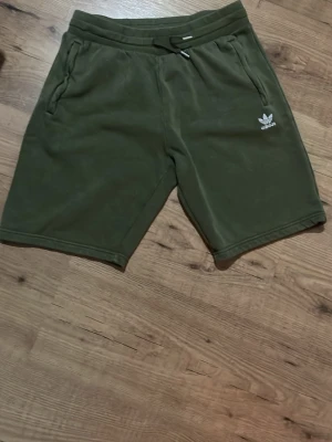 Gröna shorts från Adidas - Mörkgröna shorts från Adidas med snörning i midjan och två sidofickor. Tillverkade i mjukt material med Adidas-logga på vänster ben. Perfekta för en avslappnad stil. Stolek 170. Dom är ljusare än vad dom är på bilden 