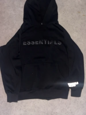 Svart Essentials hoodie med tryck - Säljer en svart hoodie från Essentials med stor logga på bröstet och mindre tryck på ärmen. Hoodien har en klassisk huva och stor magficka framtill. Perfekt för dig som gillar streetwear och enkel stil.