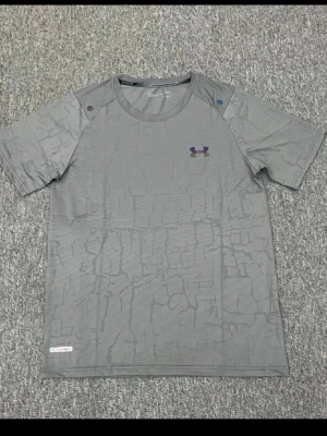 Grå mönstrad t-shirt från Under Armour - Snygg grå t-shirt från Under Armour med diskret mönster och klassisk logga på bröstet. Tillverkad i ett lätt och funktionellt material som passar perfekt till träning. Kortärmad och normal passform.