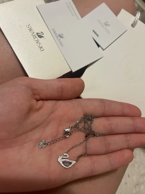 Swarovski halsband med kristaller Blå - Snyggt halsband från Swarovski i silverfärg med ett hjärtformat hänge dekorerat med gnistrande vita kristaller och en lila sten i mitten. Kedjan är tunn och elegant med justerbar längd och en liten charm vid låset.