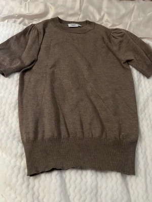 Brun stickad t-shirt från Saint Tropez - Säljer en brun stickad t-shirt från Saint Tropez i storlek XS. Tröjan har korta puffärmar, rund halsringning och ribbade muddar vid ärmslut och nederkant. Perfekt för lager-på-lager och enkel att matcha med jeans eller kjol.