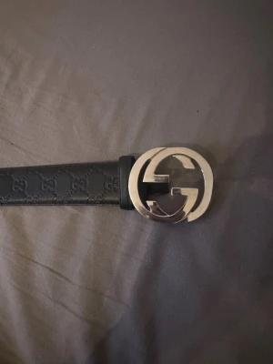 Svart gucci bälte med GG-spänne - Snyggt svart bälte med präglat GG-mönster och ett stort, rundat silverspänne i metall. Bältet har en stilren design och passar perfekt till jeans eller byxor för en trendig look. Lite slitningar vid hålen och vänster sida vid midjan. inga stora. storlek 90 med ett extra hål. 