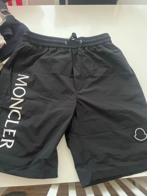 Svarta Moncler Bad shorts med snörning - Snygga svarta shorts från Moncler med vit logga längs benet och elastisk midja med snörning. Perfekta för sommaren och har en clean look med diskret Moncler-märke på ena benet. Tillverkade i lätt syntetmaterial som känns luftigt.