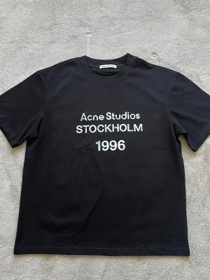 Svart Acne Studios t-shirt Stockholm - Snygg svart t-shirt från Acne Studios med vit text 'Acne Studios STOCKHOLM 1996' på bröstet. Klassisk rund hals och korta ärmar. Perfekt basplagg med stilren design och skön passform i bomull.