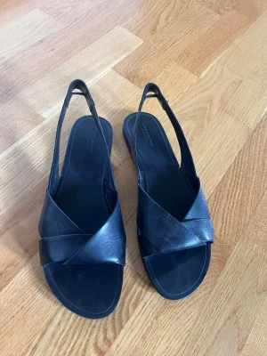 Svarta sandaler med korsade remmar - Säljer ett par stilrena svarta sandaler med breda korsade remmar och öppen tå. Sandalerna har en smal hälrem och platt sula. Tillverkade i slätt material som ger en klassisk look. Perfekta för dig som gillar minimalistisk och tidlös design.