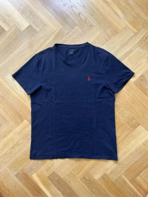 Marinblå t-shirt från Polo Ralph Lauren - Klassisk marinblå t-shirt från Polo Ralph Lauren med röd broderad logga på bröstet. T-shirten har rund halsringning och korta ärmar. Tillverkad i mjuk bomull för en skön känsla och enkel stil. En liten slitning i nacken.