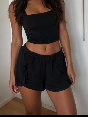 Svarta shorts med volangdetaljer strl 38 - Säljer ett par svarta shorts med elastisk midja och snygga volangdetaljer längs sidorna. De har en loose passform och är perfekta för varma dagar. Materialet känns lätt och luftigt, vilket gör dem riktigt bekväma att bära.
