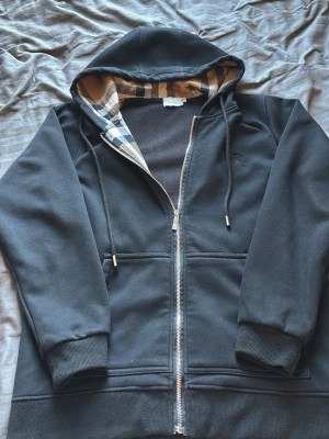 Svart hoodie med dragkedja och mönstrad huva - Svart hoodie med dragkedja och fickor framtill. Insidan av huvan har ett klassiskt rutigt mönster i beige, svart och vitt. Tröjan har ribbade muddar vid ärmslut och nederkant samt dragsko i huvan. Perfekt för en avslappnad stil.