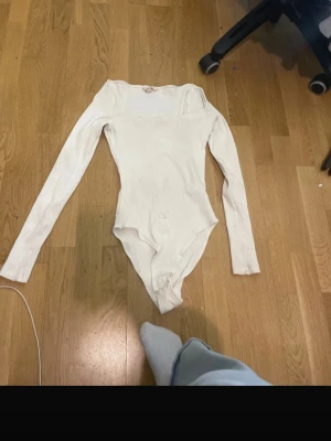 Vit ribbad body med lång ärm - Säljer en vit ribbad body med långa ärmar och rund hals. Modellen är enkel och stilren, perfekt att matcha med jeans eller kjol. Materialet är stretchigt och bekvämt, vilket gör den smidig att bära hela dagen.
